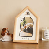 Dog house-shaped photo frame in brown wood-grain plastic with silver paw print emblem and arched white mount, displayed on a sideboard beside a dog figurine and candle.