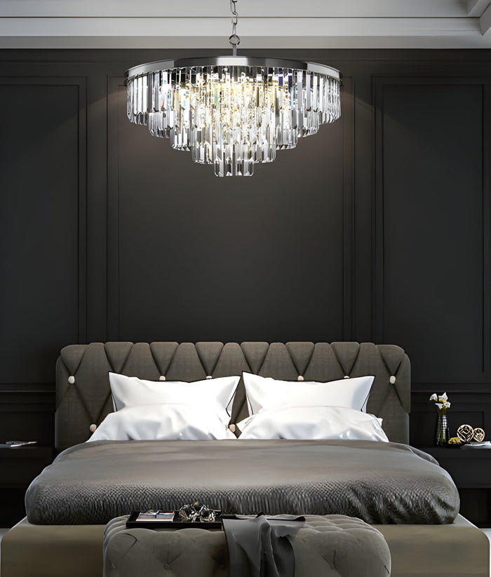 A luxurious bedroom interior featuring a plush, tufted gray headboard with white pillows and bedding. Above the bed hangs an elegant, cascading crystal chandelier. The walls are dark, adding a sophisticated ambiance.