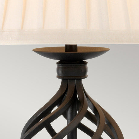 Close-up of the Belfry 1 Light Table Lamp - Black, featuring a beige pleated lampshade and a decorative black metal base with twisted design elements. This sophisticated piece highlights artisanal craftsmanship against a plain, light gray background.