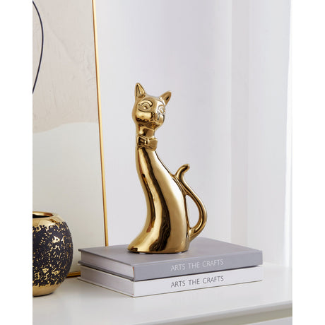 Benangin ceramic cat ornament in glossy gold with bow tie, sitting upright on a stack of books beside a gold-speckled vase in a styled room setting.