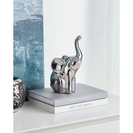 Benangin ceramic elephant ornament in mirror-like silver finish with raised trunk and fanned ears, displayed on stacked books beside a teal-patterned wall.