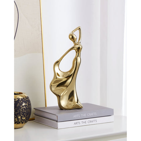 Benangin Ceramic Figurine in gold, an abstract female form with a looped raised arm and wave-like draped base, displayed on stacked books beside a dark speckled vase.