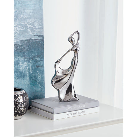 Benangin Ceramic Figurine in silver, an abstract dancing woman form with mirror-like finish and open loop body, displayed on stacked books beside a blue-grey curtain.