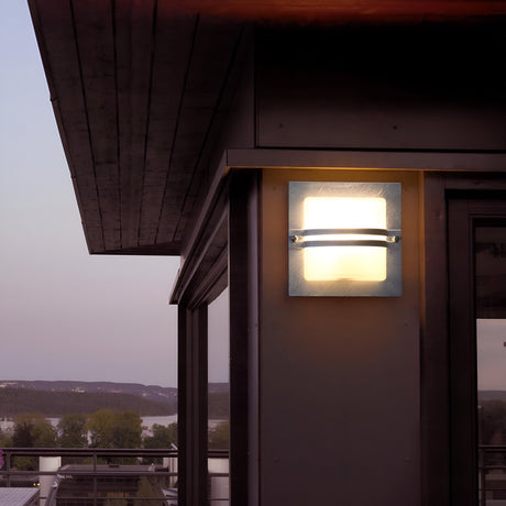 The Bern Outdoor 1 Light Wall Lantern in silver with frosted glass casts a glow on the corner of a building at dusk. The serene landscape, featuring trees and water beneath a clear evening sky, enhances the sophisticated appeal of the outdoor decor.