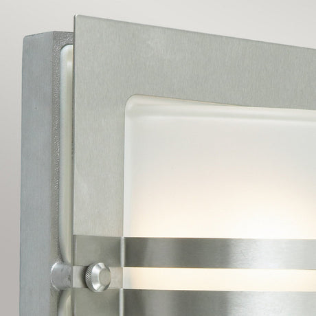 Close-up of the Bern Outdoor 1 Light Wall Lantern in stainless steel with a contemporary brushed metal finish. This fixture showcases a rectangular frosted glass panel, partially framed by metal bands, emanating a soft, warm light.