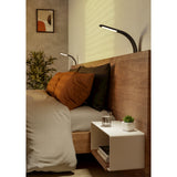 Bevadoro Clamp Lamp in black with curved arm and flat LED head, clamped to a wooden bed headboard above pillows and a white nightstand.