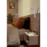 Bevadoro LED clamp lamps in white with chrome head caps, mounted on a wall-fixed headboard above a bed, illuminating a bedroom with a bedside shelf and plant.