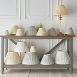 Bevdon 20cm tapered lamp shade in ivory pleated silk, shown among an assortment of pleated lamp shades on a console table and lit table lamp.