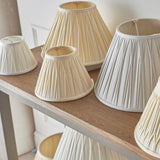 Bevdon 20cm tapered lamp shade in ivory silk with box pleat design, shown among various pleated shades of different sizes and finishes on a wooden shelf.