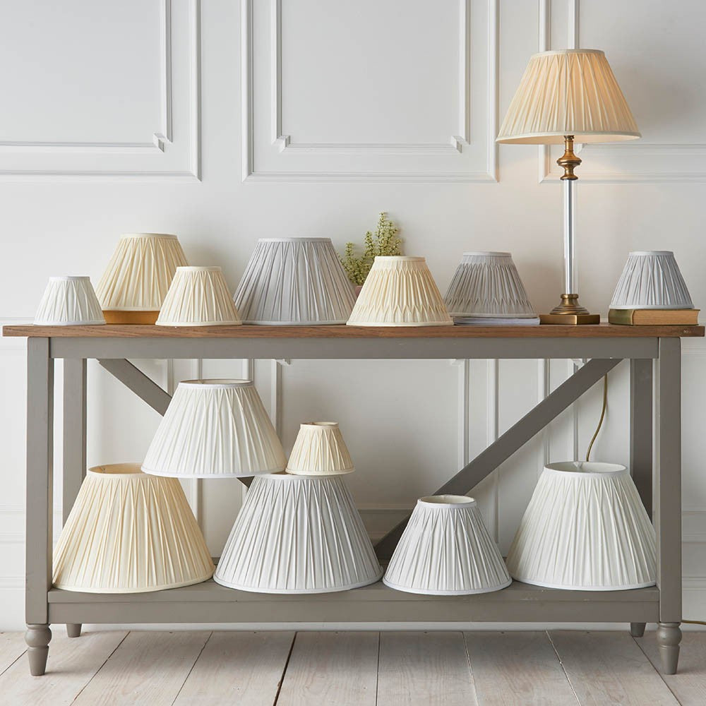 Endon Bevdon 25cm tapered ivory silk lamp shades with pinch pleat detail, displayed among assorted shades on a grey console table against a panelled wall.