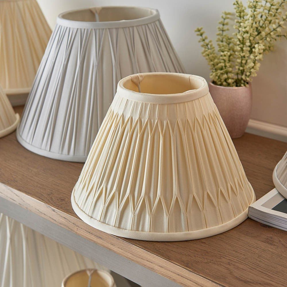 Endon Bevdon 25cm tapered lamp shade in ivory silk with hand-stitched double pinch pleats, displayed on a wooden shelf beside a grey pleated shade.