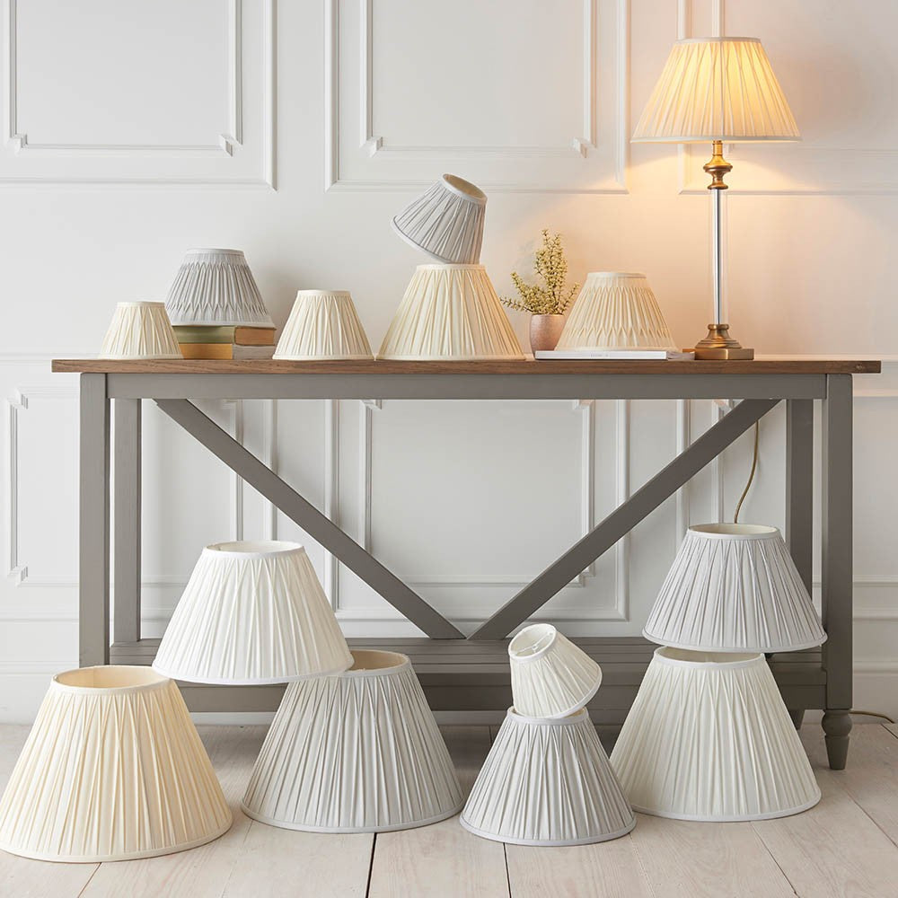 Endon Bevdon 25cm tapered lamp shade in ivory silk with pinch pleats, displayed among a collection of pleated shades on and around a grey console table.