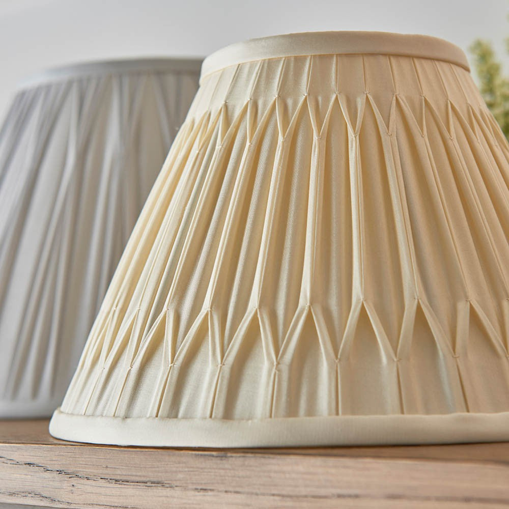 Endon Bevdon 25cm tapered lamp shade in ivory silk with hand-stitched pinch pleats, shown beside a grey pleated shade on a wooden surface.