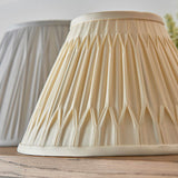 Endon Bevdon 25cm tapered lamp shade in ivory silk with hand-stitched pinch pleats, shown beside a grey pleated shade on a wooden surface.