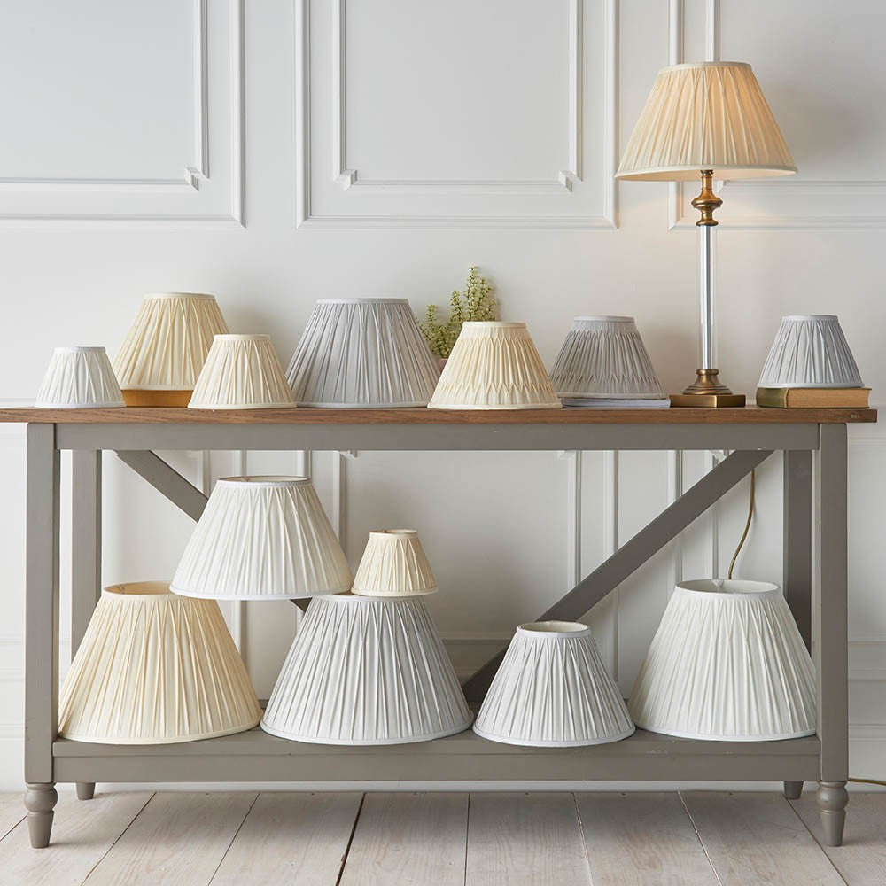 Endon Bevdon tapered lamp shades in ivory silk with pinch pleats, displayed in various sizes on a grey console table alongside a lit lamp.