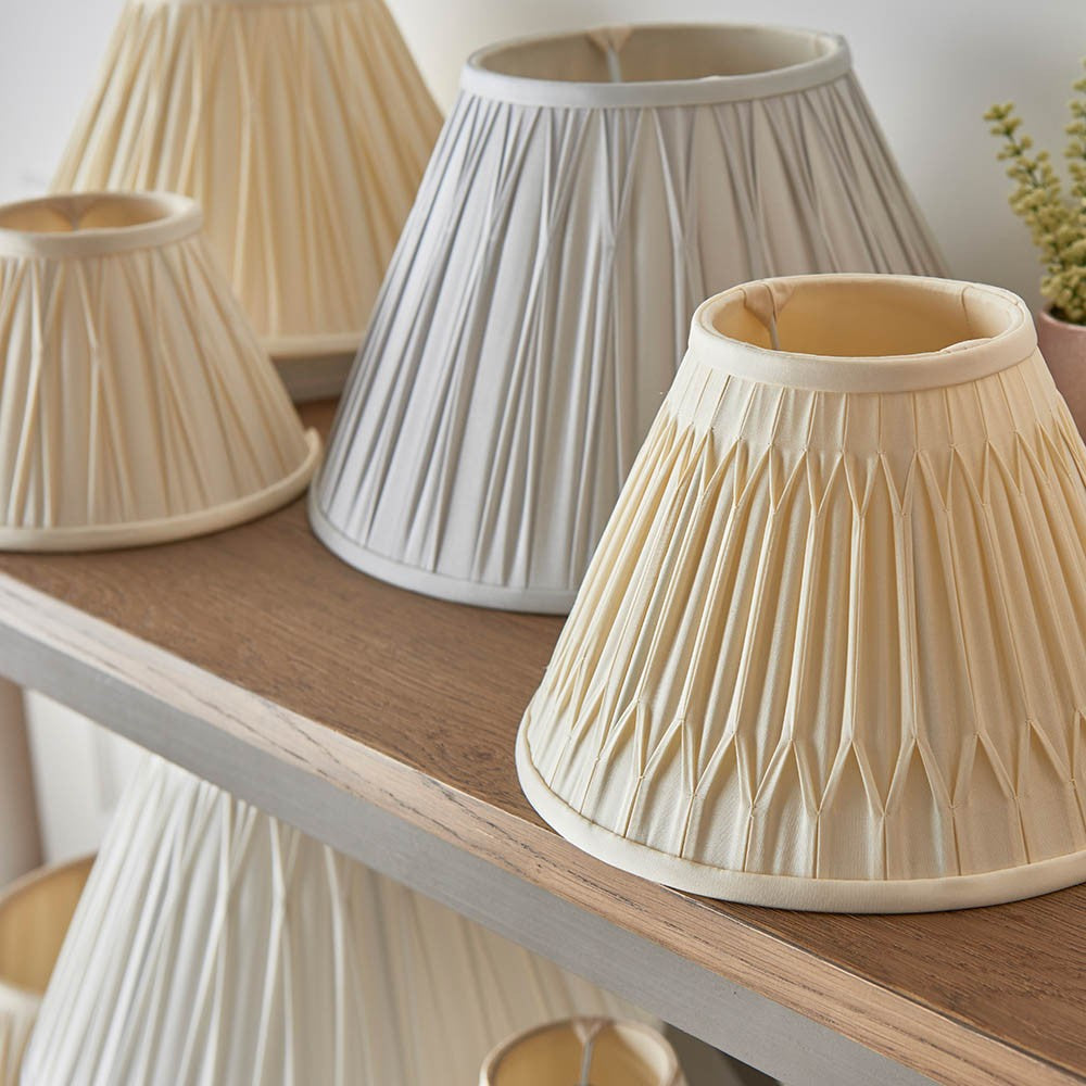Endon Bevdon 30cm tapered lamp shade in ivory silk with hand-stitched pinch pleats, displayed on a wooden shelf alongside other pleated shades.