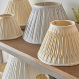 Endon Bevdon 30cm tapered lamp shade in ivory silk with hand-stitched pinch pleats, displayed on a wooden shelf alongside other pleated shades.