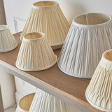 Bevdon 35cm tapered lamp shade in ivory silk with box pleat design, shown among various pleated shades in cream and grey on a wooden shelf.