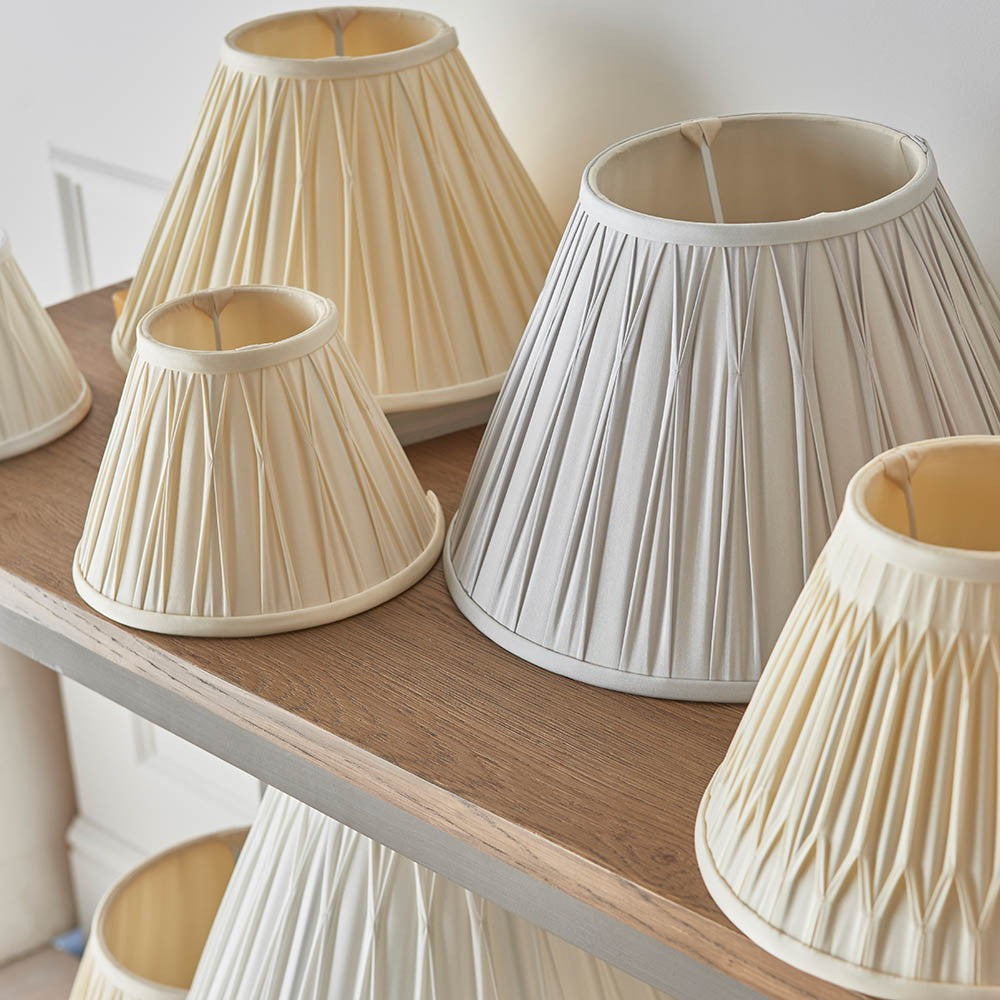 Endon Bevdon tapered lamp shades in ivory silk with pinch-pleat detail, displayed in various sizes on a wooden shelf against a pale wall.
