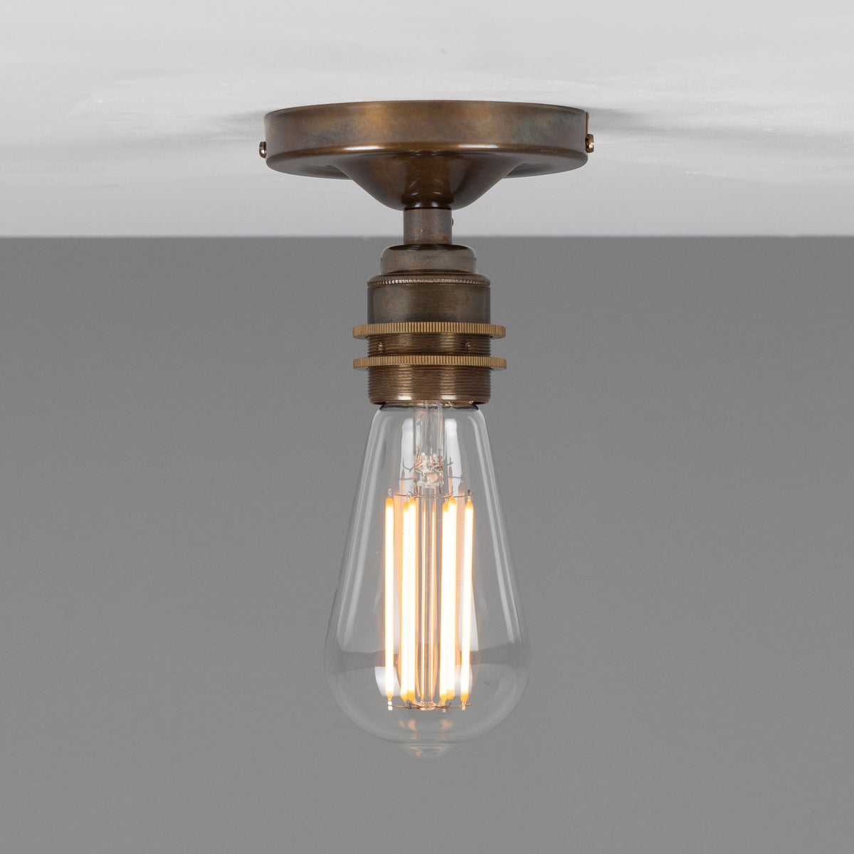 Bexter Vintage Brass Flush Ceiling Light with round ceiling plate and single knurled lampholder holding an exposed filament bulb, shown against a plain grey background.