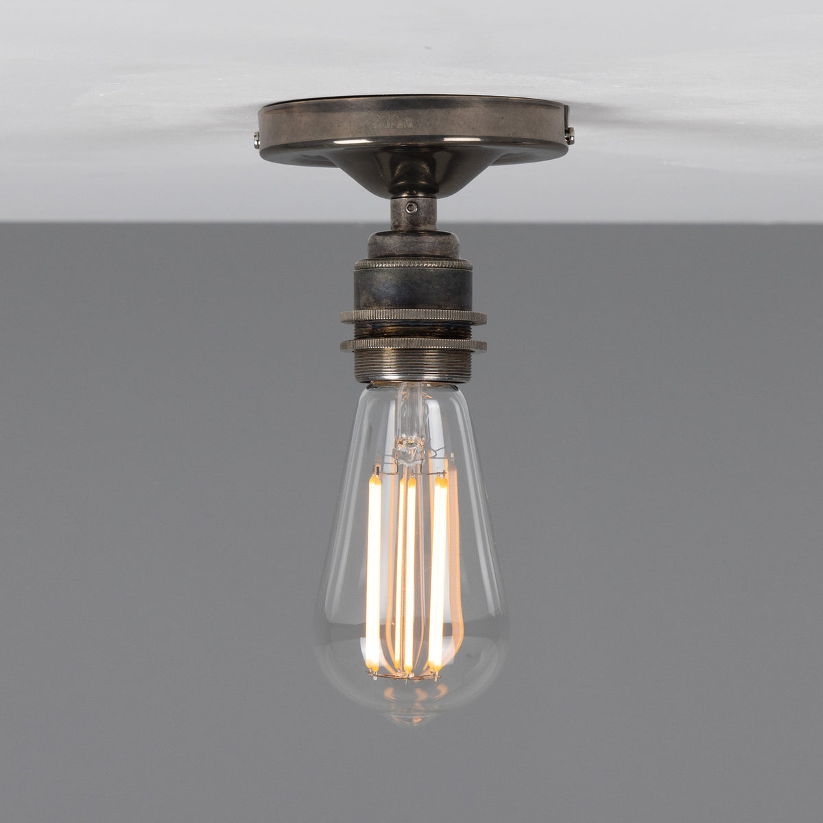Bexter Vintage Brass Flush Ceiling Light with round ceiling plate and single knurled lampholder holding an exposed bulb, shown against a plain grey background.