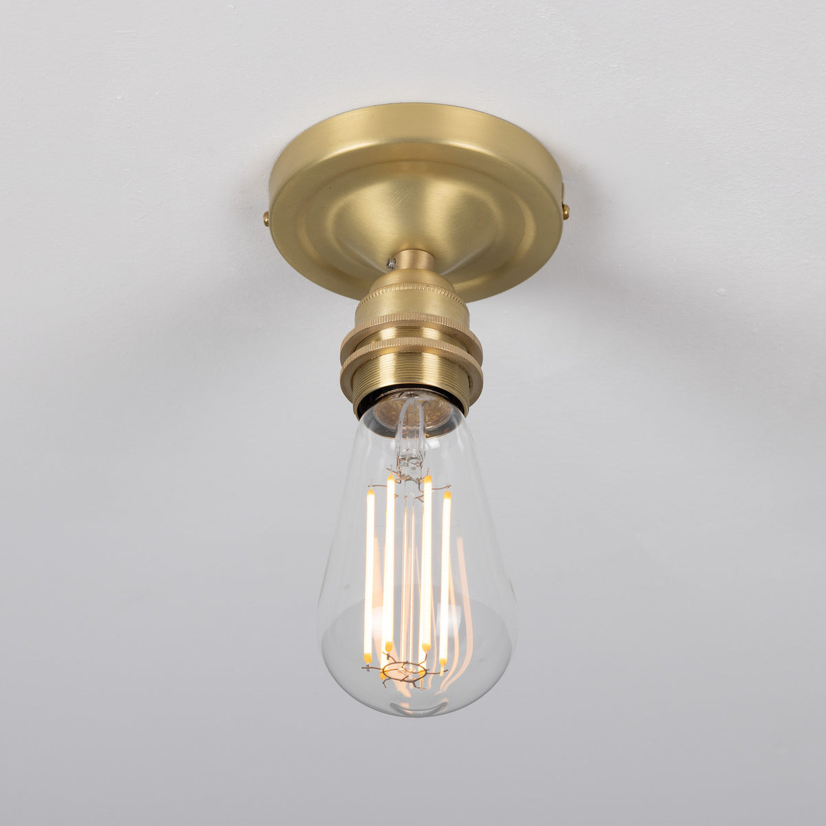 Bexter Vintage Brass Flush Ceiling Light with round ceiling plate and single knurled lampholder holding a bare filament bulb, shown against a plain white background.