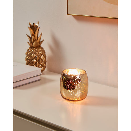 Bezamby glass tealight holder in gold with a rounded, mirror-finish body and silvery speckles, lit on a shelf beside a gold pineapple ornament.