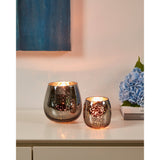 Bezamby glass tealight holders in smoky grey with mirrored, speckled finish, shown lit in two sizes on a sideboard beside books and blue hydrangeas.