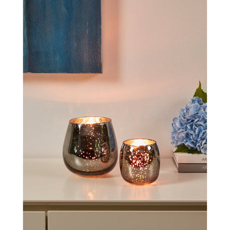 Bezamby glass tealight holders in smoky grey with mirrored, speckled finish, shown lit in two sizes on a sideboard beside books and blue hydrangeas.