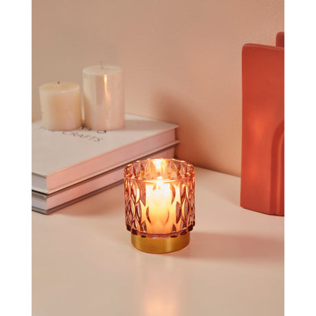 Bezamby pink glass tealight holder with faceted sides and gold-coloured base, lit on a table beside pillar candles, books, and a terracotta vase.