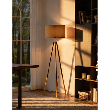 Bidford Floor Lamp in brown with a tripod wood and steel base and mocha fabric drum shade, shown lit in a living room beside a bookshelf.