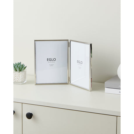 Bitata Double Photo Frame in polished silver metal, hinged book-style design with two glass-fronted portrait panels, shown open on a sideboard beside a small plant and books.