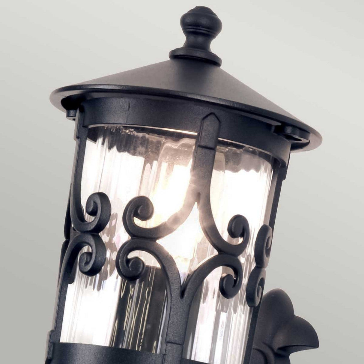 A close-up of the Hereford 1 Light Outdoor Wall Lantern in black showcases its intricate scrollwork and glass enclosure. This lantern-style design is beautifully enhanced by a warm glow from within, casting a soothing ambiance through textured glass against a neutral backdrop.