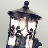 A close-up of the Hereford 1 Light Outdoor Wall Lantern in black showcases its intricate scrollwork and glass enclosure. This lantern-style design is beautifully enhanced by a warm glow from within, casting a soothing ambiance through textured glass against a neutral backdrop.
