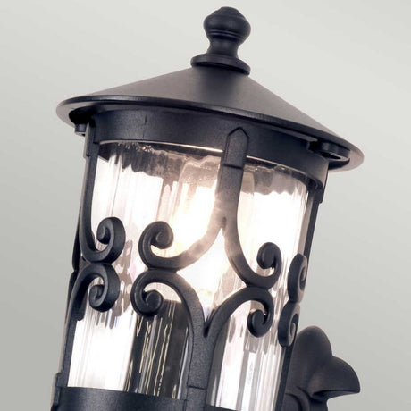 A close-up of the Hereford 1 Light Outdoor Wall Lantern in black showcases its intricate scrollwork and glass enclosure. This lantern-style design is beautifully enhanced by a warm glow from within, casting a soothing ambiance through textured glass against a neutral backdrop.