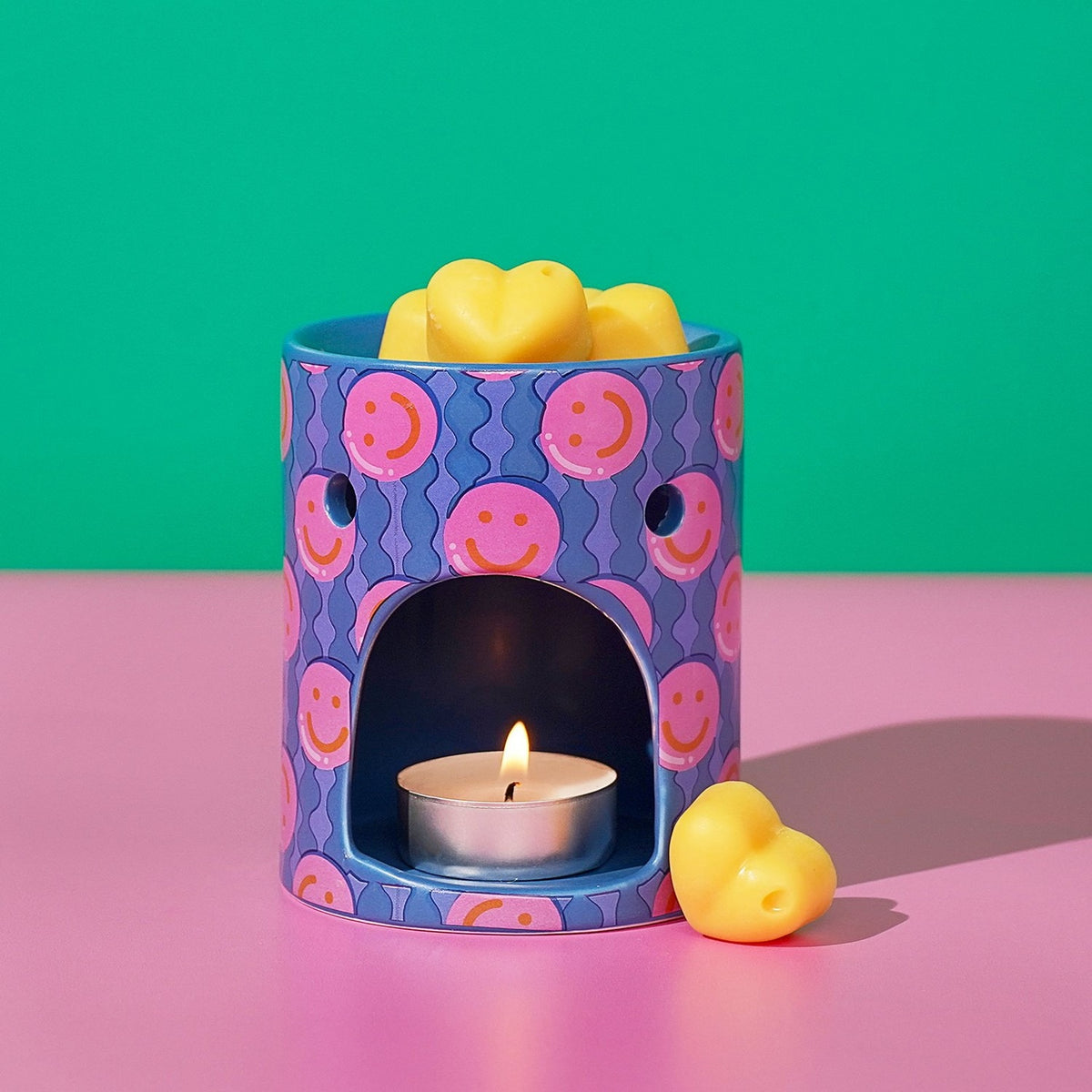 Round ceramic wax melt burner in blue with pink smiley face print, lit tealight visible through arched opening, yellow heart-shaped wax melts on top and beside it.