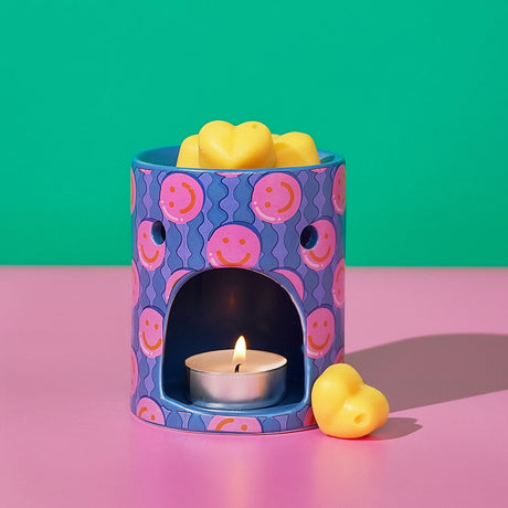 Round ceramic wax melt burner in blue with pink smiley face print, lit tealight visible through arched opening, yellow heart-shaped wax melts on top and beside it.