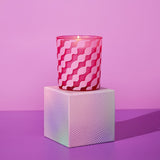 Dream Big Wavy Stripe Round Scented Candle in pink glass with diagonal wavy stripe pattern, lit wick, displayed on a patterned pedestal against a purple background.