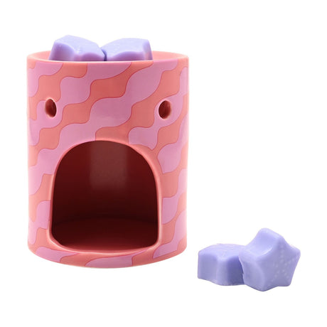 Round ceramic wax melt burner in pink with wavy coral stripes, arched tealight opening, and lilac star-shaped wax melts on top and beside it on a white background.