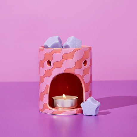 Round ceramic wax melt burner in pink with wavy coral stripes, arched tealight opening, lit candle inside, and lilac star-shaped wax melts on top and beside it.