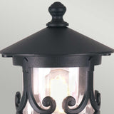 The Hereford 1 Light Pedestal Lantern - Black showcases a black lantern-style with intricate scrollwork, complete with a clear glass casing that reveals the bulb inside, all set against a light gray background.