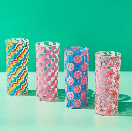 Set of 4 tall glass tumblers, 290ml, each with a different print: rainbow diagonal wavy stripes, pink wavy stripes, pink smiley faces on blue, and pink daisies, shown on a green background.