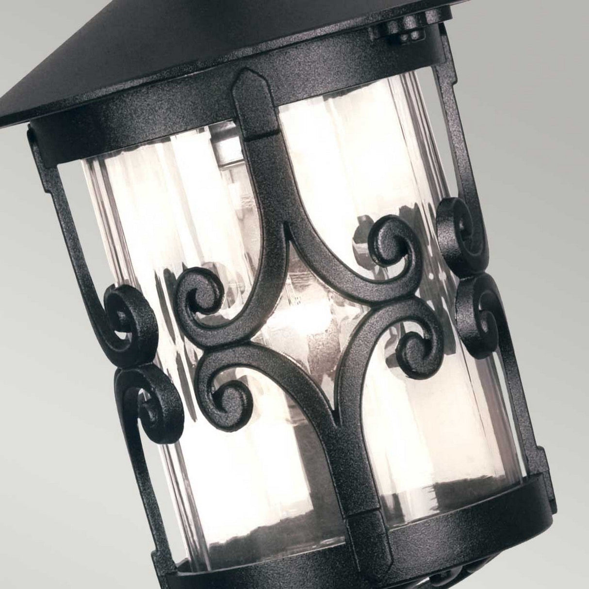 Close-up of the Hereford 1 Light Outdoor Porch Chain Pendant in black, showcasing its traditional design with intricate scrollwork on glass panels. Tilted at an angle, the visible light bulb makes it perfect for outdoor lighting. The background is a simple, light gray.