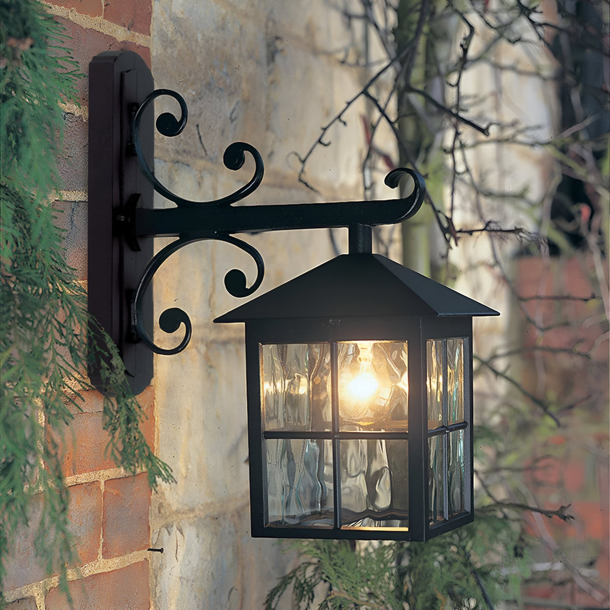 The Winchester-2 1 Light Outdoor Wall Down Light - Black, made from die-cast aluminium, is mounted on a brick wall. This vintage lighting fixture emits a warm glow through its glass panels, nestled amid green foliage and a stone wall.