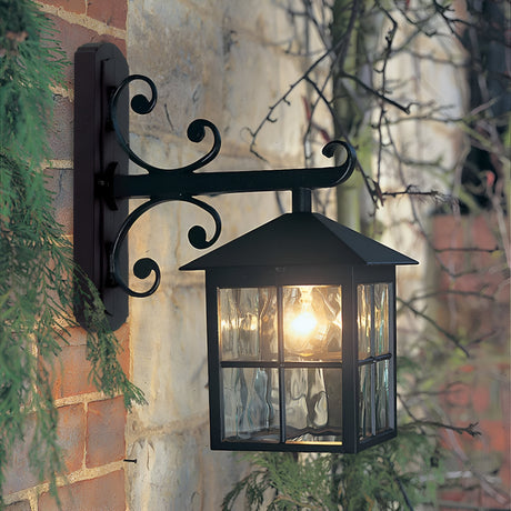 The Winchester-2 1 Light Outdoor Wall Down Light - Black, made from die-cast aluminium, is mounted on a brick wall. This vintage lighting fixture emits a warm glow through its glass panels, nestled amid green foliage and a stone wall.