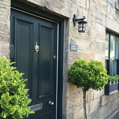 A quaint stone cottage facade features a dark green door with a brass knocker, flanked by neatly trimmed topiary bushes. Above the window box filled with plants, the charming Hereford 1 Light Outdoor Wall Down Lantern in black completes the look. A sign reads West Cottage.