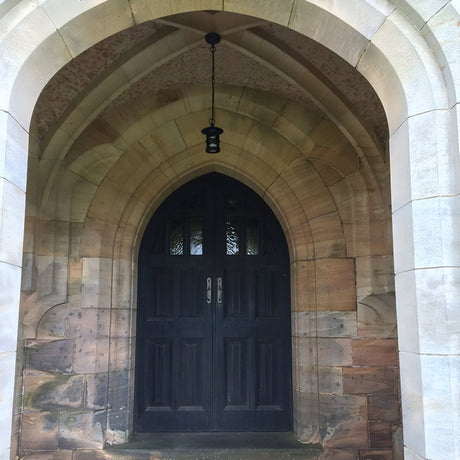 A dark wooden door with arched stonework surrounds in a Gothic architectural style is complemented by the Hereford 1 Light Outdoor Chain Pendant in black overhead. The outdoor lighting highlights the textured, multicolored stone, casting an enchanting glow on its classic lantern shape.