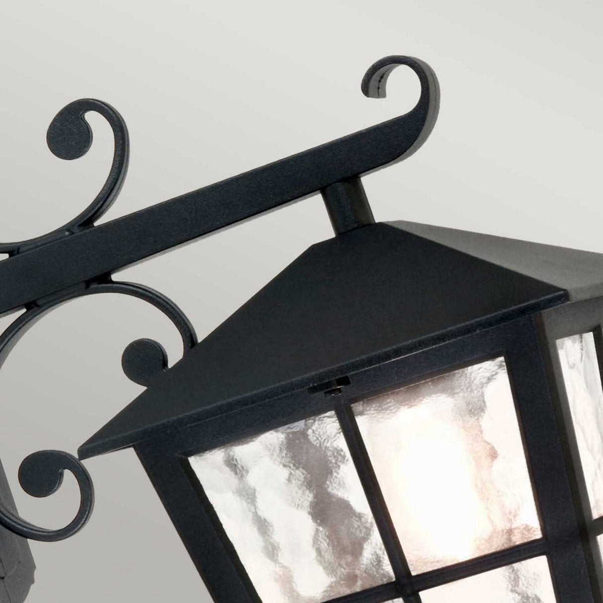 The Canterbury Outdoor Wall Lantern Down - Black showcases a close-up view of a lantern made from durable die-cast aluminum. Its glass panels allow a warm glow to illuminate the intricate scrollwork details.