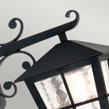 The Canterbury Outdoor Wall Lantern Down - Black showcases a close-up view of a lantern made from durable die-cast aluminum. Its glass panels allow a warm glow to illuminate the intricate scrollwork details.