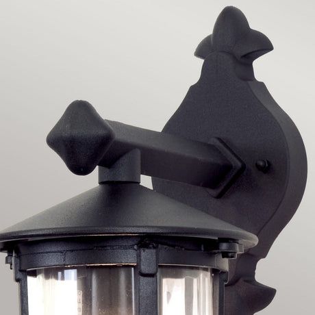 The Hereford Outdoor Wall Down Lantern - Black is a classic outdoor wall fixture styled like a lantern. It is made with a decorative mount from die-cast aluminium, featuring a transparent glass cover and topped with a pointed finial, all beautifully contrasted against its light grey backdrop.